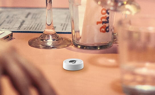A white Flic Message Button on a restaurant tabletop, surrounded by glassware and a menu.