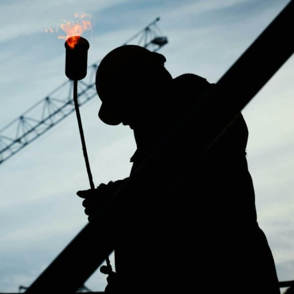 Silhouette of a hardhatted worker holding a flare against a twilight sky.