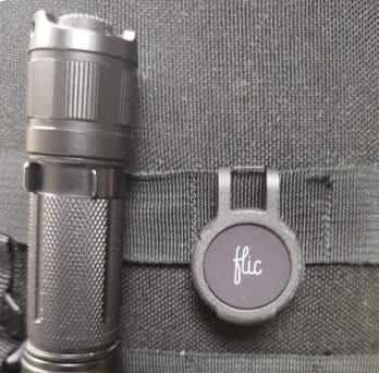 A black Flic button with a protective bumper, clipped to a tactical equipment vest in field-ready gear.